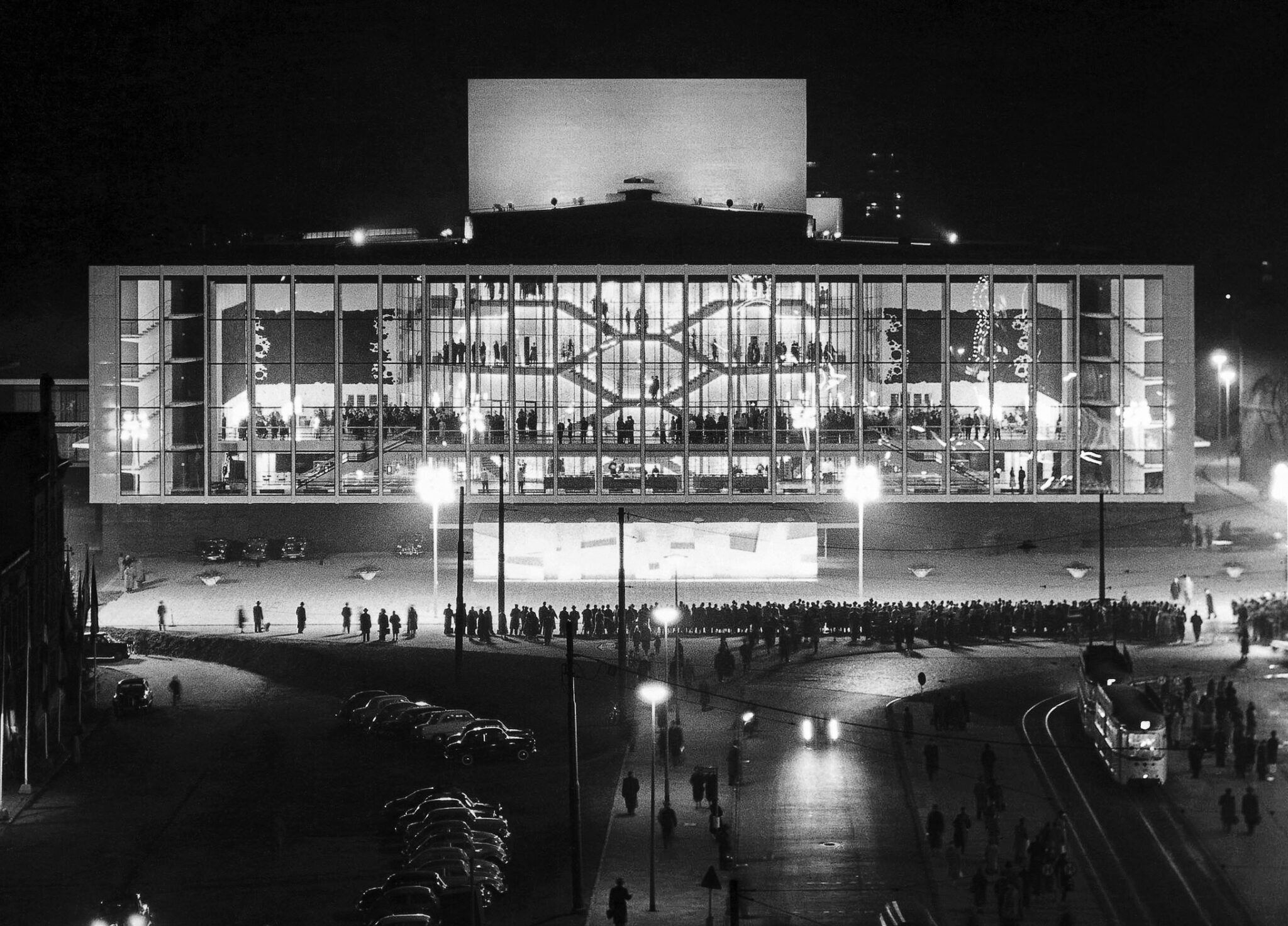 Nachtaufnahme des Großen Hauses am Abend der Eröffnung, 15.Dezember 1959 mit Bürgern auf dem Vorplatz: Bei Lichtgleichgewicht ist das Glas transparent, der Theaterbesucher wird von außen gesehen zum Akteur und das Theaterfoyer zum wettergeschützten Bereich des öffentlichen Raumes -